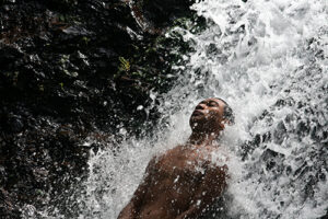 A barechested Samoan man leans back into a gushing waterfall with black rock in the background