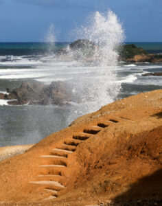 A staircase cut into red sea cliff in Dominica. In the background, you can see waves smashing spray into the air, boulders, sea and blue sky. The image is very dynamic