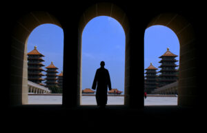 Three arches and a monk silhouetted against a bright blue sky. Pagodas with red roofs flank all sides of the square the monk is walking into